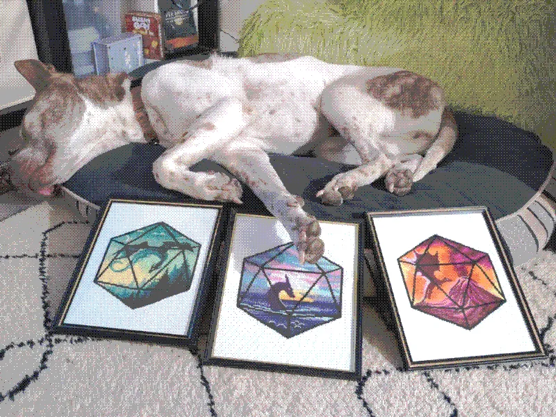 3 framed cross stitches of dragons, one blue, one red, and one green, layed in front of my dog sleeping on a dog bed